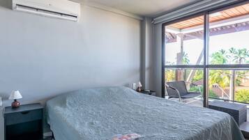 A bedroom with a bed, a nightstand, a lamp, and a window with a view of palm trees.