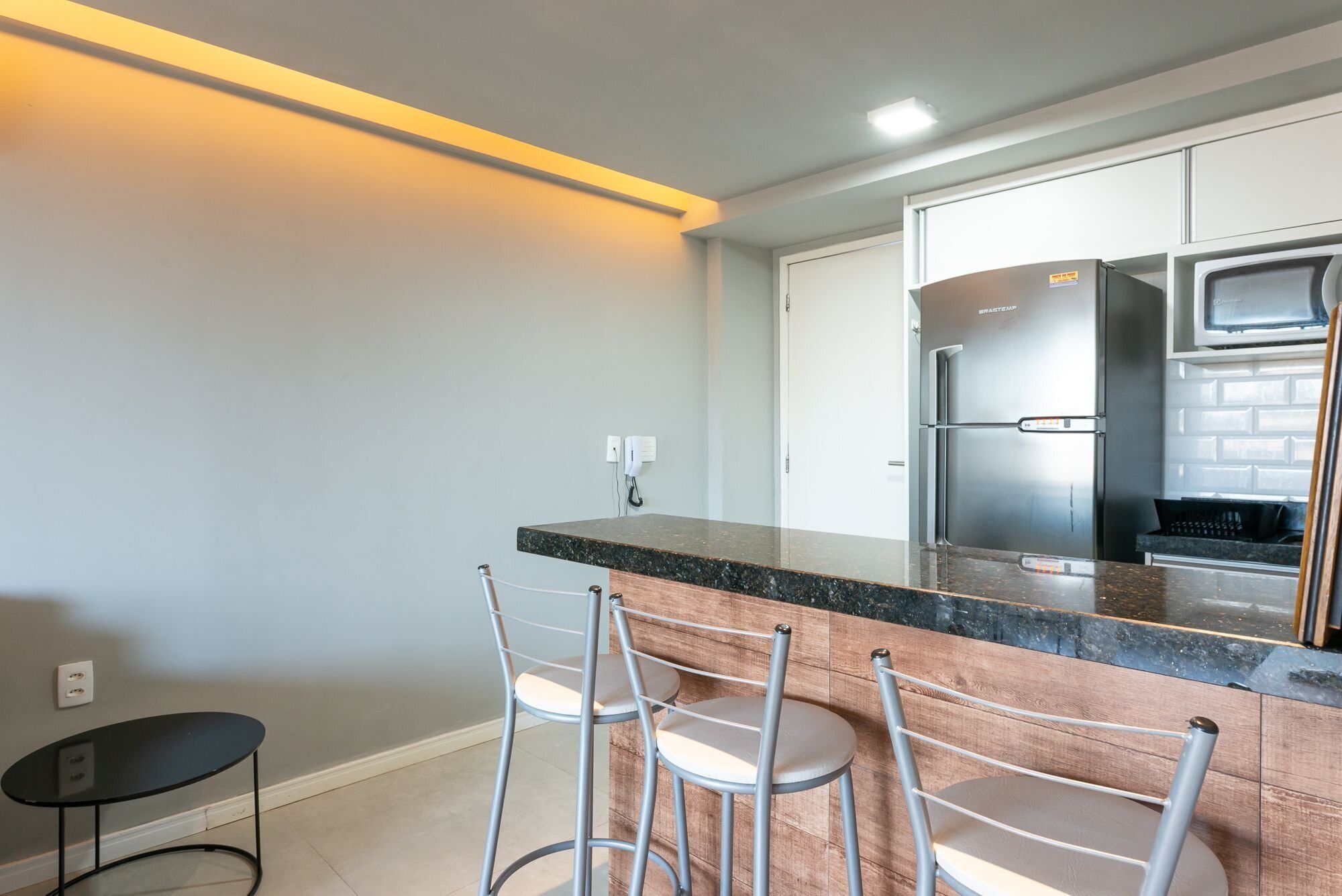 A modern kitchen with a stainless steel refrigerator, a granite countertop, and three bar stools.