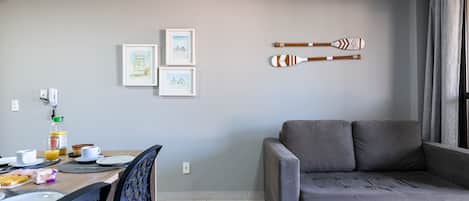 A modern living room with a grey sofa, a dining area with a table set for breakfast, and two framed artworks on the wall.