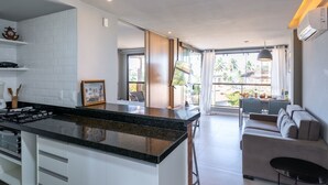 A modern kitchen with a central island, a dining area with a sofa and table, and a view of palm trees through large windows. - Praia Central Porto de Galinhas (Ipojuca)