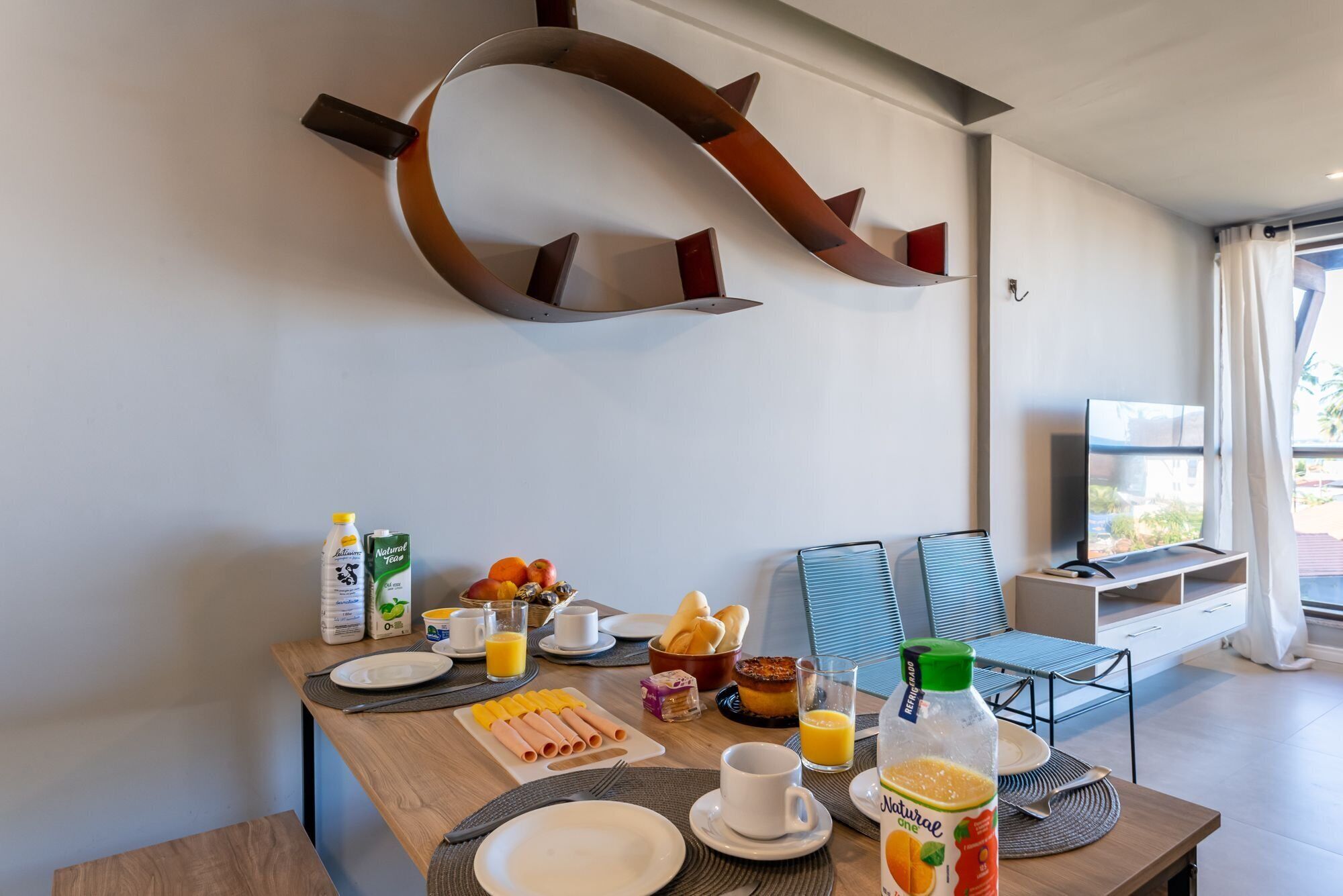 A breakfast setup with a variety of food items and beverages on a wooden table, a TV in the background, and a modern wall design.