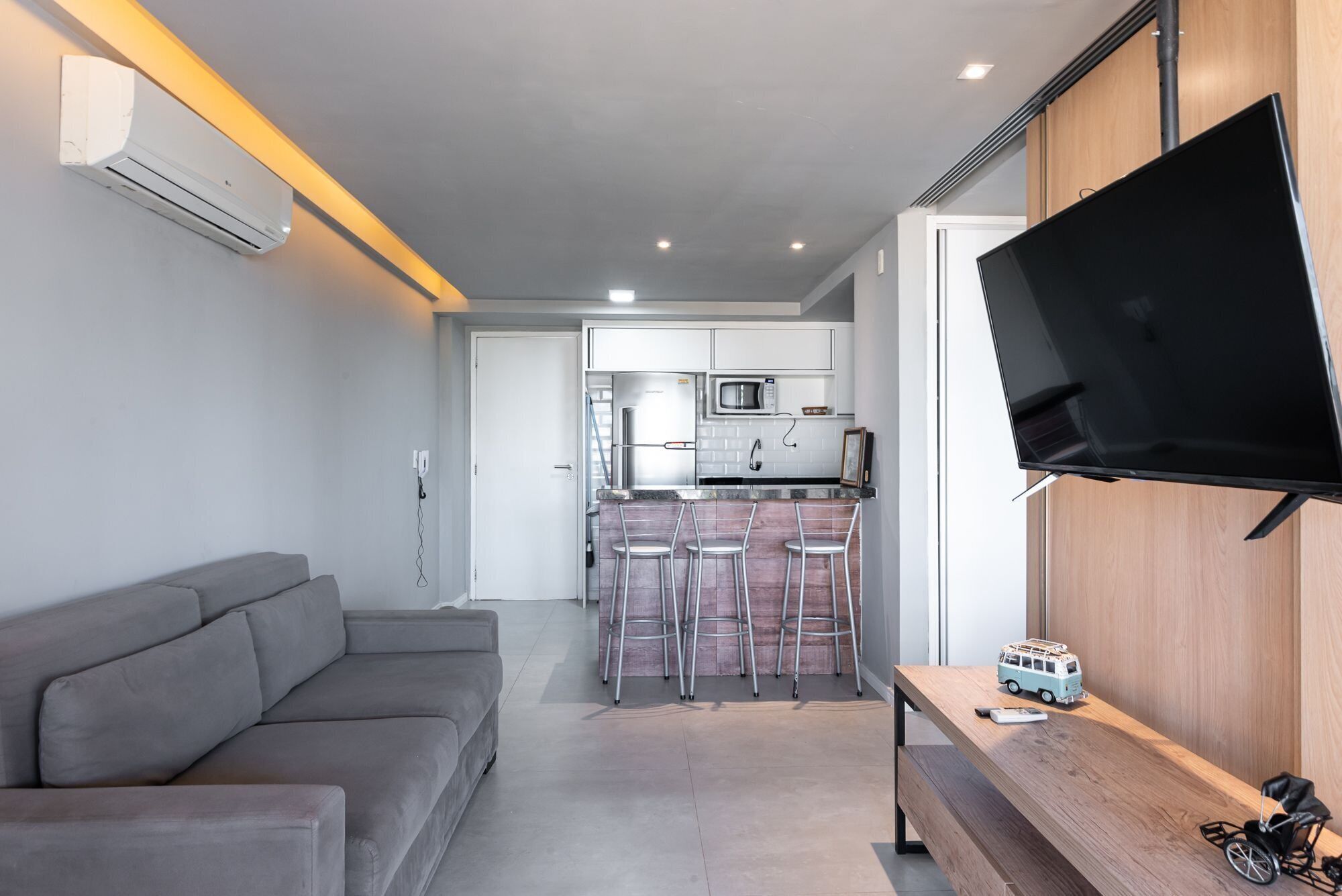Un salon moderne avec un canapé gris, une table à manger en bois et un téléviseur fixé au mur, et une cuisine en arrière-plan.