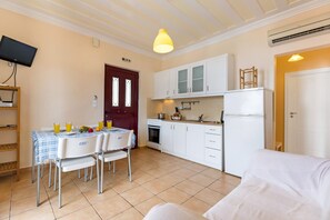 Une cuisine de taille réduite avec des meubles blancs, une table à manger avec des chaises, un téléviseur fixé au mur et un ventilateur de plafond.