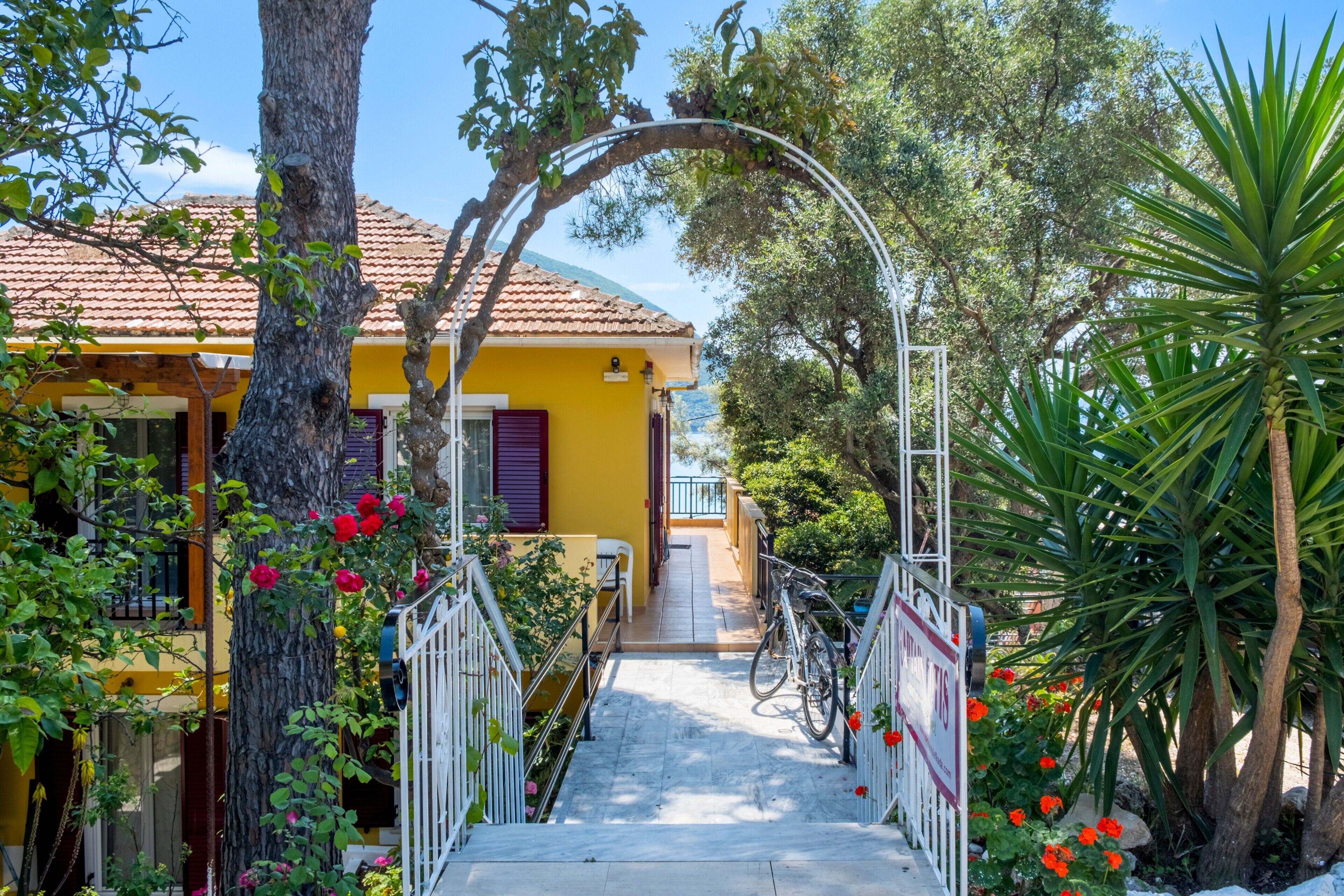 A yellow building with a staircase leading to a balcony, surrounded by trees and plants.
