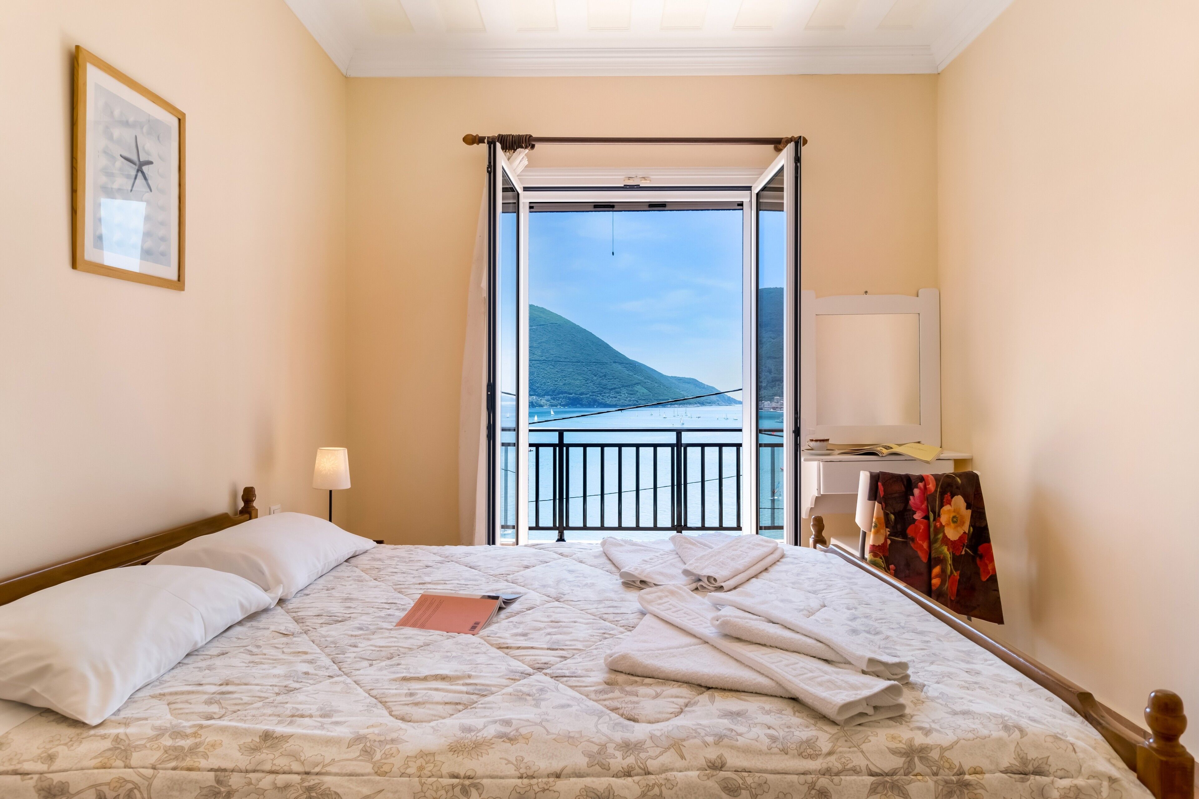 Une chambre d’hôtel comprenant un lit, une lampe de chevet, un tableau encadré, une fenêtre donnant sur une vue de montagne et de l’eau, et un balcon avec une balustrade.
