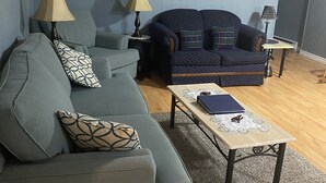 Un salon avec un canapé gris, un canapé bleu à motifs, deux lampes, une petite table avec un livre et un élément décoratif mural.