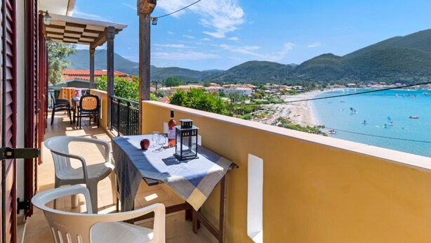 Un balcon avec une table et des chaises, donnant sur une plage où l’on aperçoit des bateaux et un paysage montagneux.