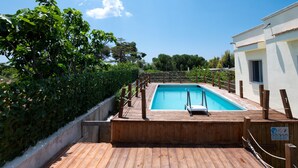 Une terrasse en bois avec une piscine, entourée de verdure et bordée d’un bâtiment blanc.
