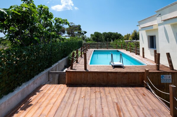 A wooden deck with a swimming pool, surrounded by greenery and a white building.
