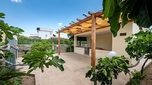 Un patio extérieur aménagé, abrité par une pergola, doté d’un sol en pierre et entouré de verdure.