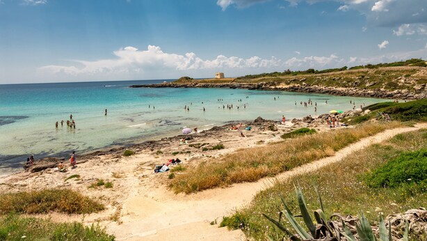 Une plage à l’eau turquoise et limpide, où l’on peut voir des gens nager et prendre des bains de soleil, et un bâtiment situé au loin, sur un promontoire rocheux.