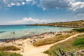 Ein Strand mit klarem türkisblauem Wasser, Menschen beim Schwimmen und Sonnenbaden, und ein entferntes Gebäude auf einem Felsen.
