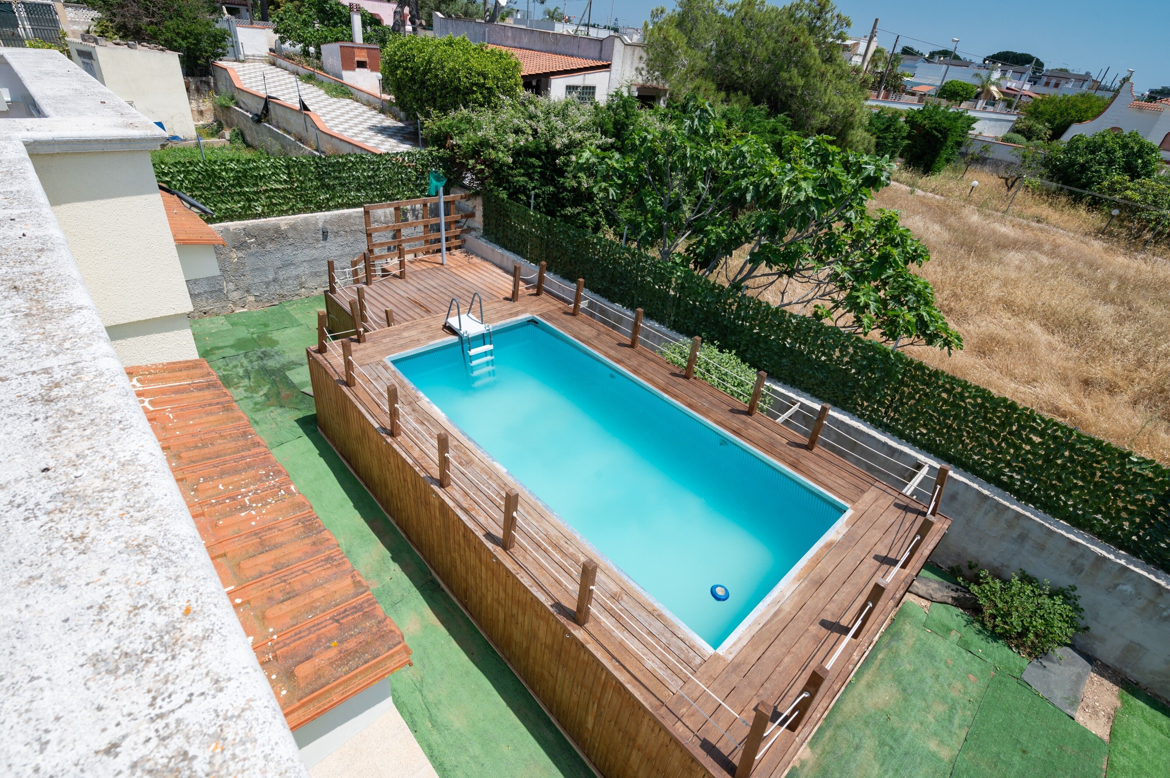 A rectangular pool with a wooden deck and a ladder, surrounded by greenery and residential buildings.