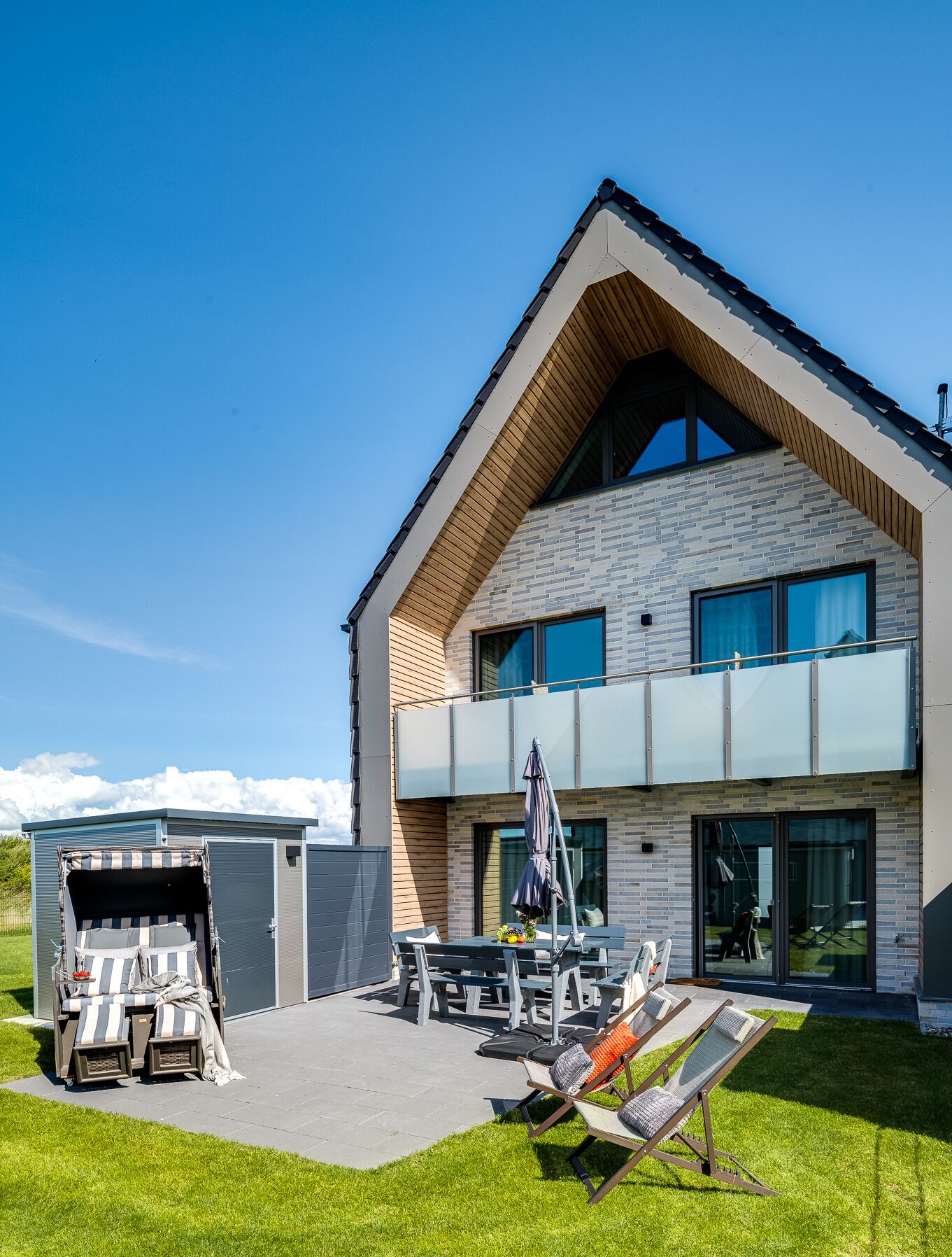 Une maison moderne à deux étages, dotée d’un toit en pente, d’un patio couvert aménagé avec du mobilier d’extérieur et d’un petit garage attenant.