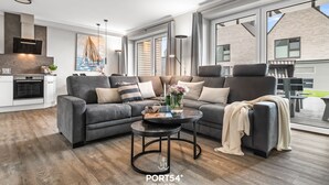 A modern living room with a sectional sofa, a round coffee table, and a large window offering a view of the outside. - Momme II - close to the North Sea and the beach, sauna, terrace, modern (Büsum)