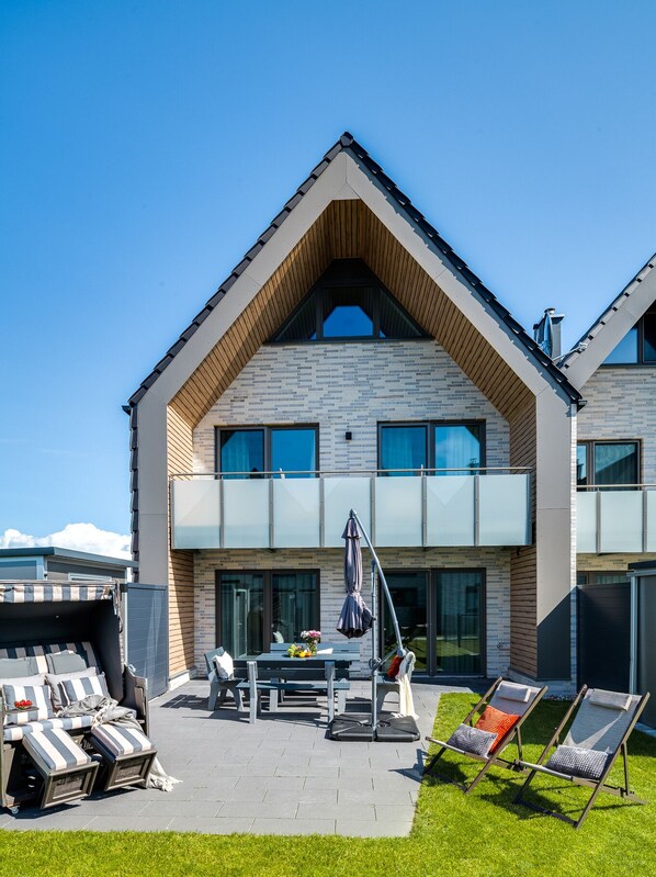A modern two-story house with a sloped roof, large windows, and a patio area with outdoor furniture.