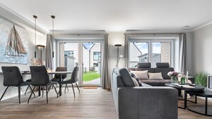 A modern living room with a grey sofa, dining area with chairs, and a large window offering a view of the outdoors. - Momme II - close to the North Sea and the beach, sauna, terrace, modern (Büsum)