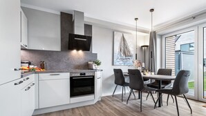 A modern kitchen with white cabinets, a built-in oven, and a dining area with a table and chairs. - Momme II - close to the North Sea and the beach, sauna, terrace, modern (Büsum)