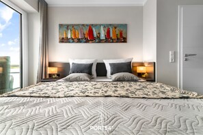 A hotel room with a large bed, two bedside tables, a painting of sailboats, and a view of the outdoors.