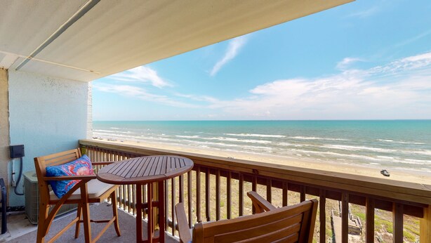 Un balcon avec une table en bois et deux chaises, donnant sur une plage, l’océan et un ciel dégagé.