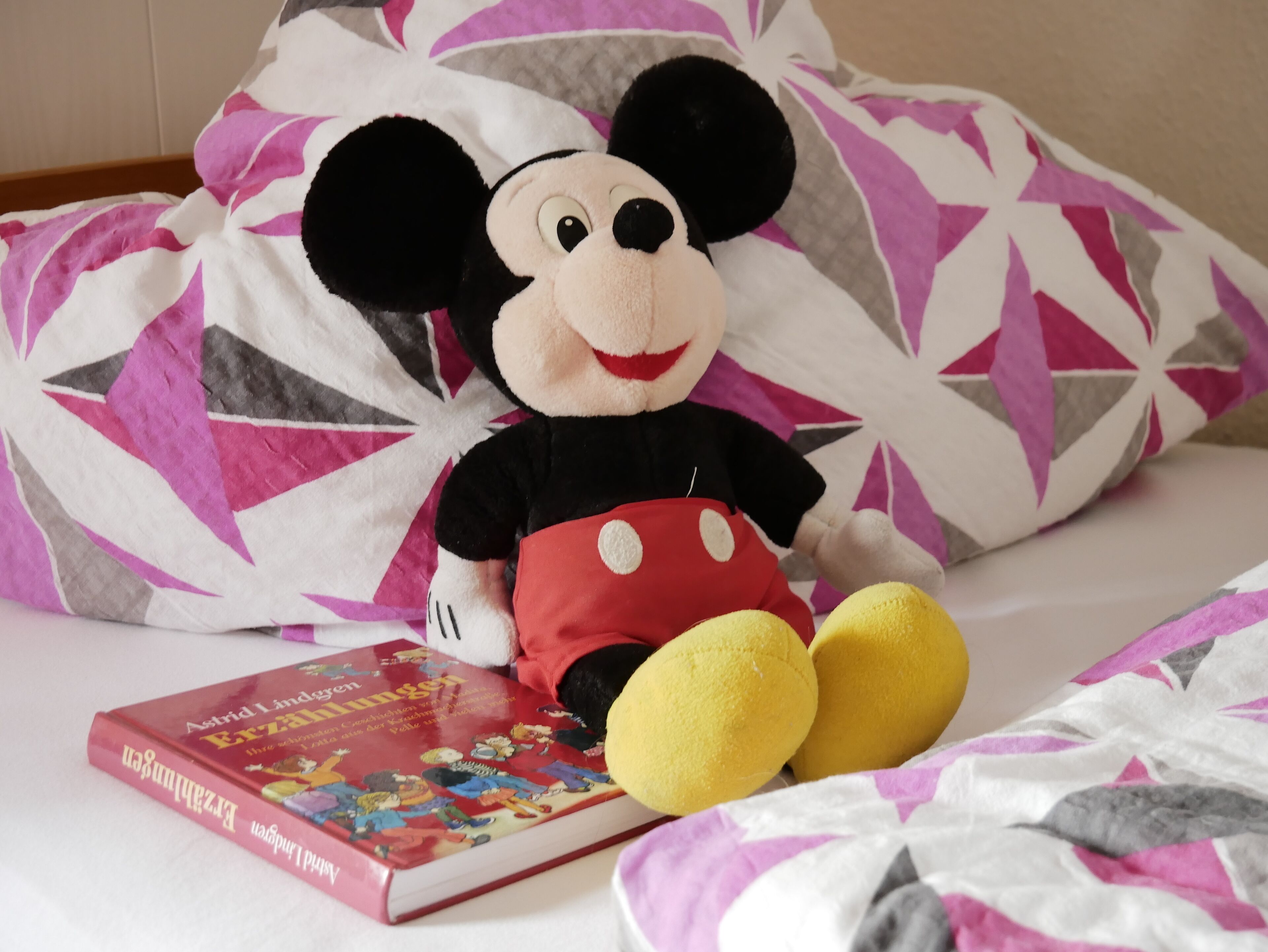 Un jouet en peluche est posé sur un lit, à côté d’un oreiller à motifs et d’un livre.