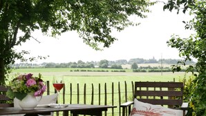 Eine Holzbank mit Kissen, ein Tisch mit Blumen in einer Vase und einem Glas Rotwein, und ein Blick auf ein grünes Feld durch einen Holzzaun.