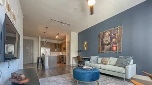 A modern living room with a grey sofa, a blue ottoman, and a patterned rug. A kitchen is visible in the background. - Boulevard Stays Dashing 1BR Suite Pool, Parking Gym G (Richardson)