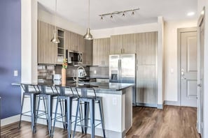 A modern kitchen with a central island, stainless steel appliances, and bar stools. - Boulevard Stays Dashing 1BR Suite Pool, Parking Gym I (Richardson)