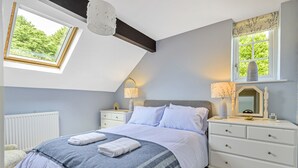 Un dormitorio con tragaluz, una cama con manta azul y blanca a rayas, una mesita de noche con lámpara y un aparador con cajones.