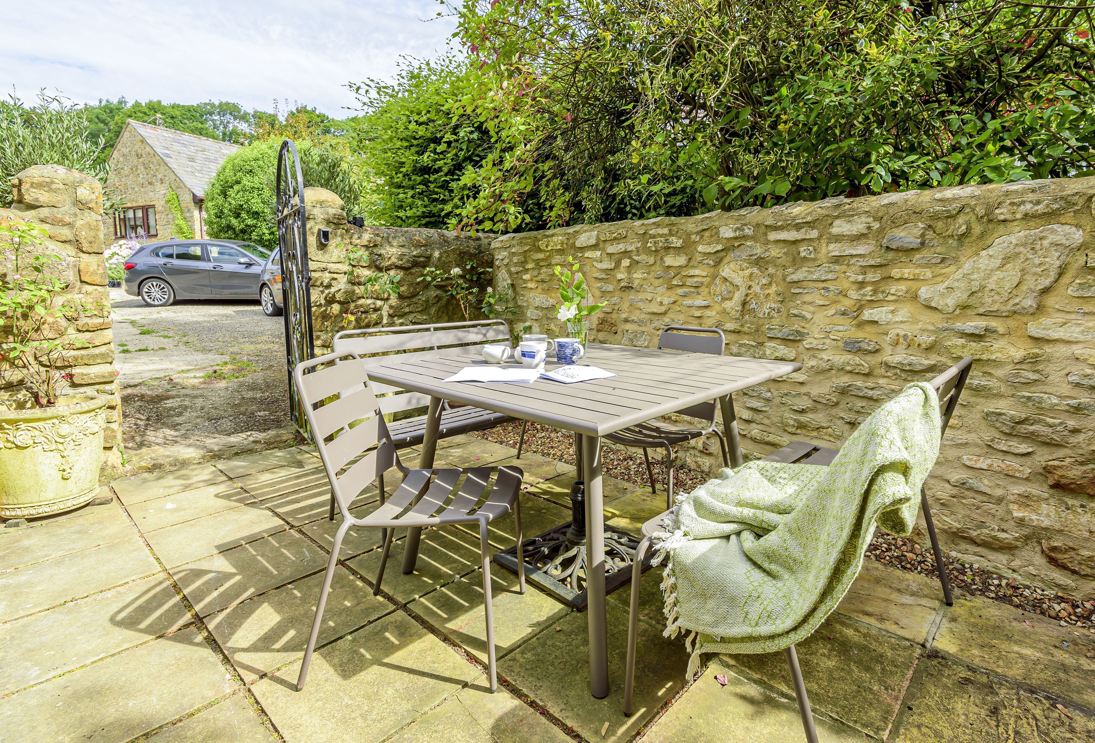 Eine Terrasse im Freien mit Tisch und Stühlen, eine Steinmauer und ein geparktes Auto im Hintergrund.