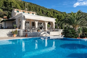 Un espace aménagé au bord de la piscine, avec un bâtiment doté de fenêtres en arc de cercle et d’un patio couvert.