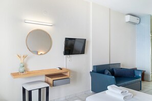 Une chambre d’hôtel moderne équipée d’un téléviseur à écran plat, d’un miroir rond, d’un bureau en bois avec un tiroir, d’un canapé bleu et d’un lit avec une serviette pliée.