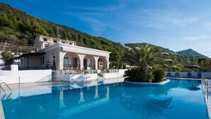 A swimming pool with a building featuring arched windows and a terrace, surrounded by greenery and hills. - Apartment in a Hotel 'Sunset Premium Studio 3' with Sea View, Shared Pool, & Wi-Fi (Lefkada)