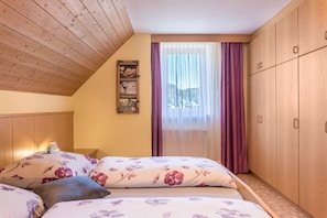 Ein Schlafzimmer mit schrägverdachter Decke, einem Holzschrank, einem Bett mit floralem Bettzeug, einem Nachttisch und einem Fenster mit durchsichtigen Vorhängen.