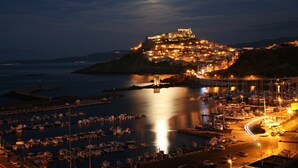A coastal town at night with a prominent castle-like structure on a hill, a marina with boats, and a bridge connecting to the mainland. - Affittimoderni Castelsardo La Gemma (Castelsardo)