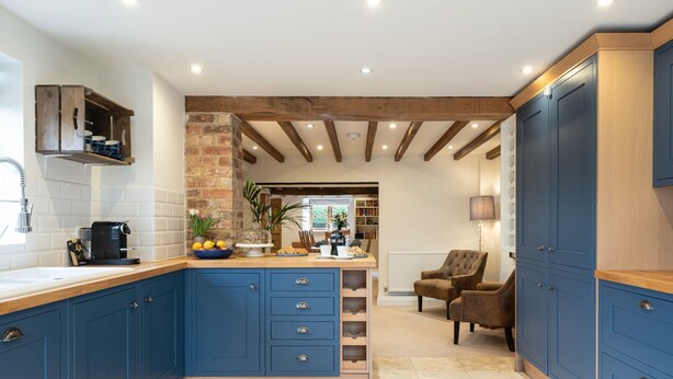 A kitchen with blue cabinets, a brick wall, and a wooden ceiling. - Family friendly period holiday cottage in the Cotswolds - The Old Post Office (Ilmington, near Chipping Campden)