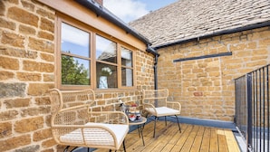 A wooden deck with wicker chairs, a small table, and a window overlooking greenery. - Family friendly period holiday cottage in the Cotswolds - The Old Post Office (Ilmington, near Chipping Campden)