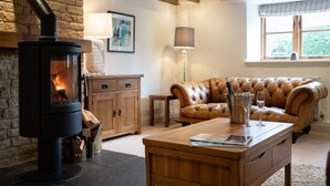 A cozy living room with a stone fireplace, leather sofa, wooden coffee table, and a window with a view. - Family friendly period holiday cottage in the Cotswolds - The Old Post Office (Ilmington, near Chipping Campden)