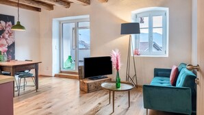 Ein Wohnzimmer mit Holzboden, einer Couch, einem Fernseher auf einem Holzschrank, einer grünen Vase mit rosa Blumen und einer Lampe.