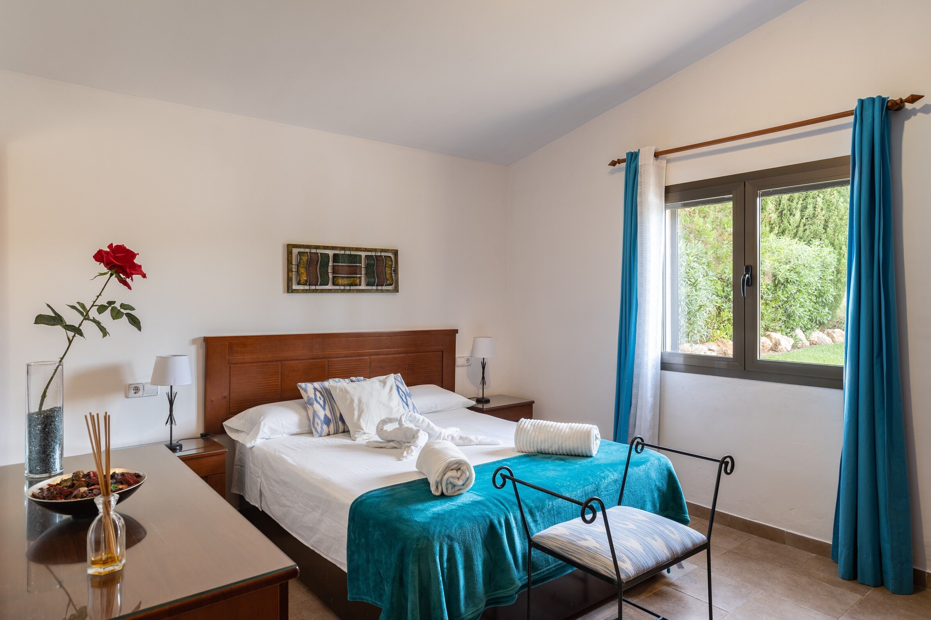 Une chambre d’hôtel avec un lit, des tables de chevet, une fenêtre avec des rideaux, un vase avec une fleur et un tableau au mur.