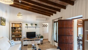 A living room with a sofa, TV, wooden beams, and a large wooden door. - Villa Can Tomas with Mountain View, Garden, Pool & Wi-Fi (Pollença)