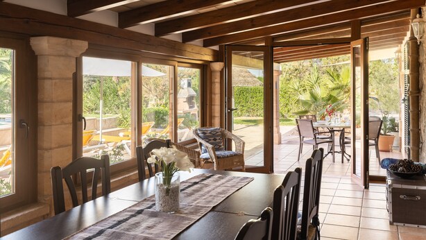 A dining area with a table set for four, surrounded by chairs, with a view of the garden and outdoor seating. - Villa Can Tomas with Mountain View, Garden, Pool & Wi-Fi (Pollença)