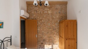 A bedroom with a stone wall, a bed with white linens, a wooden wardrobe, and a ceiling light fixture. - Villa Can Tomas with Mountain View, Garden, Pool & Wi-Fi (Pollença)