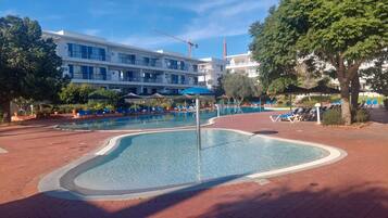 Una piscina dalla forma particolare, circondata da lettini e ombrelloni, con un edificio a più piani sullo sfondo.