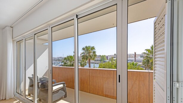 Un balcón con vistas a palmeras y edificios, una puerta corrediza de vidrio y un área de descanso con una mesa y sillas.