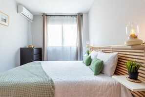Une chambre à coucher avec un lit, une table de chevet avec une lampe, une plante en pot, une commode et une fenêtre avec des rideaux.