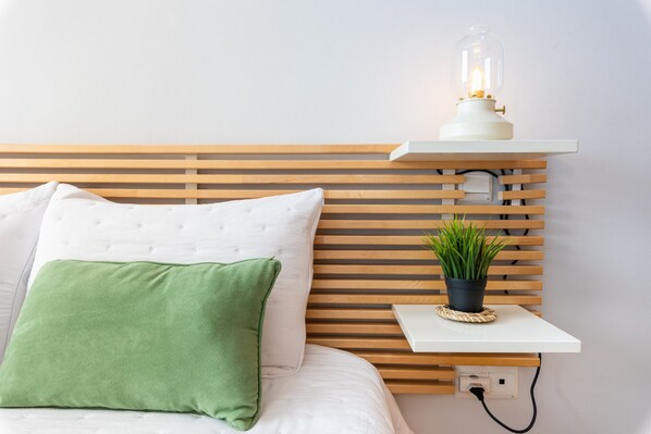 Ein ordentlich bezogenes Bett mit einem grünen Kissen, einem weißen Kissen und einem weißen Kopfteil mit horizontalen Lamellen. Darauf befindet sich ein kleiner Tisch mit einem Blumentopf und einer Lampe.