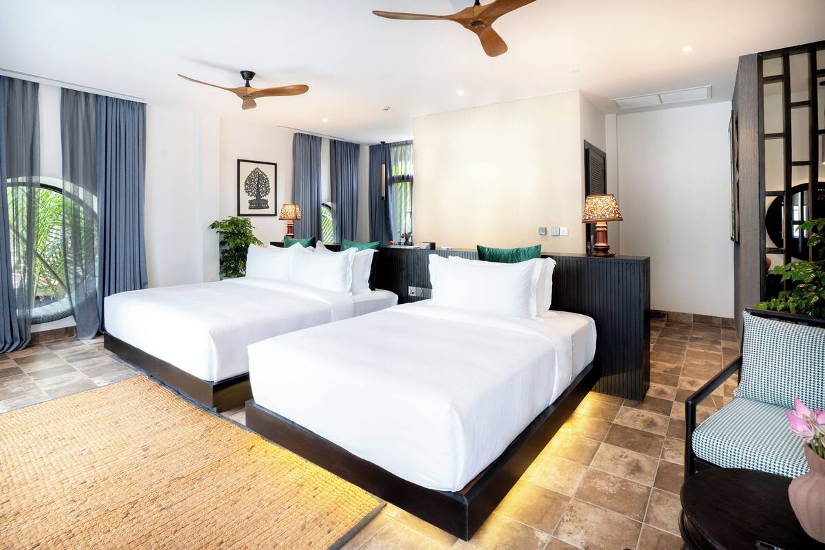 A hotel room with two beds, a ceiling fan, a desk with a lamp, and a view of greenery outside.