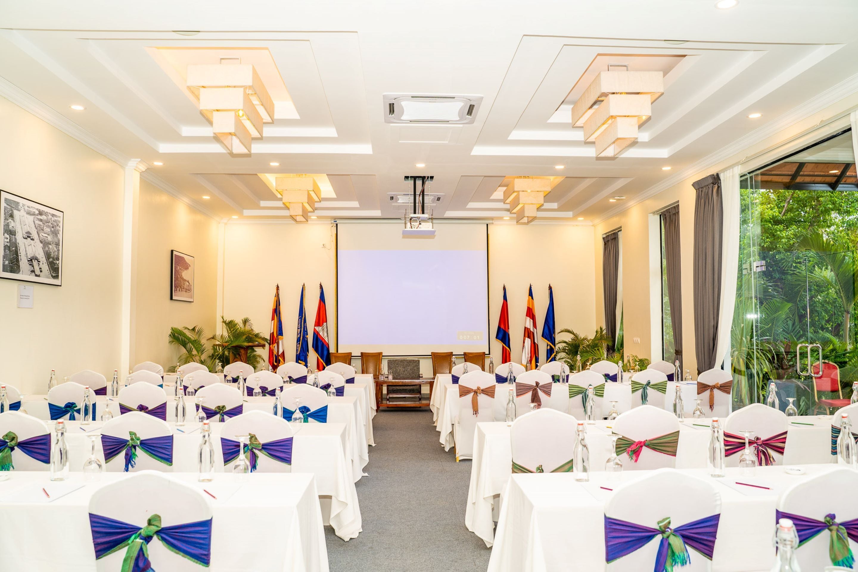 A conference room with rows of tables, chairs with bow ties, a projector screen, and flags.