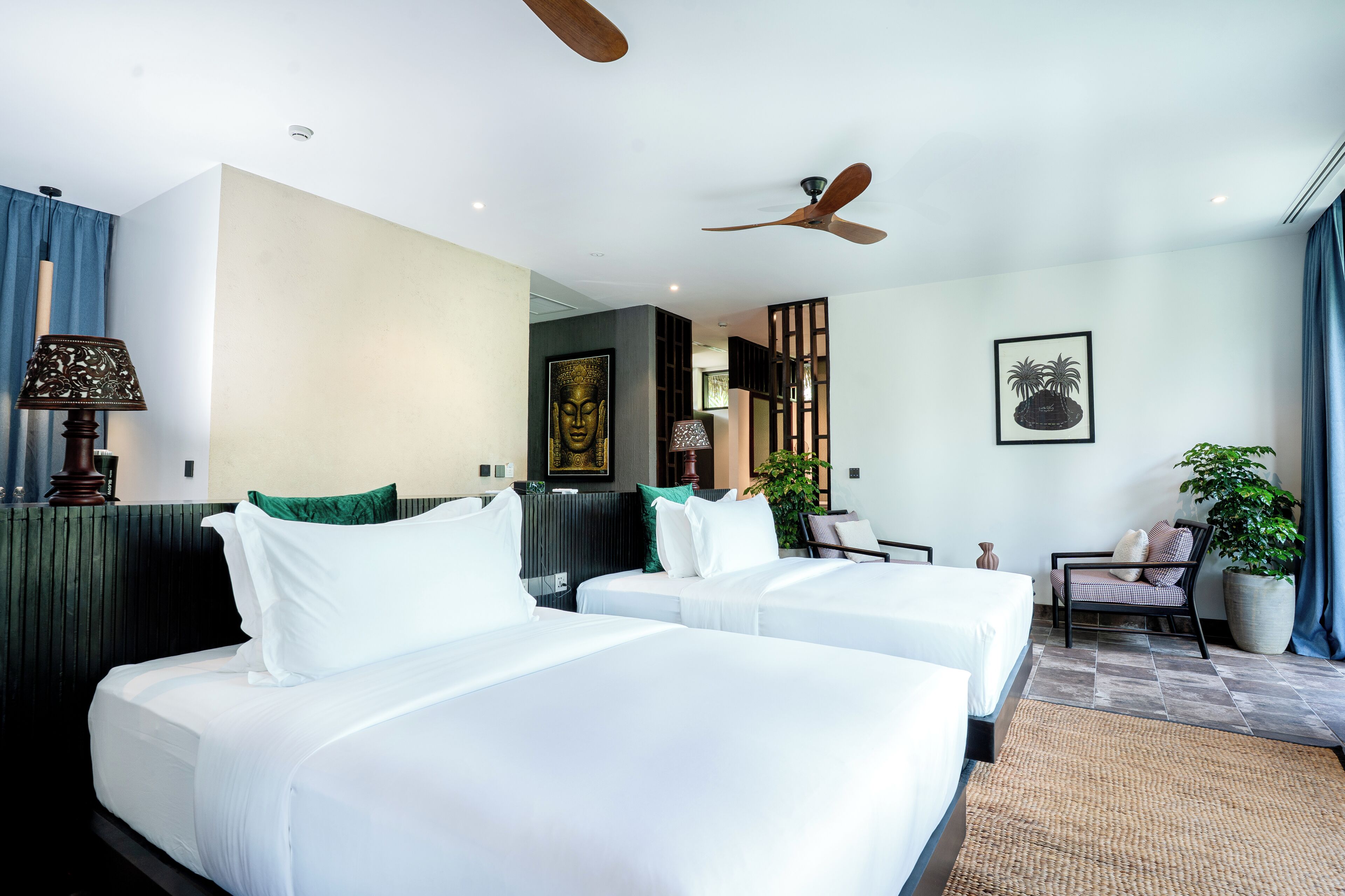 Une chambre d’hôtel avec deux lits, un ventilateur de plafond, une grande plante en pot et une chaise.